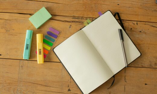 Top view of opened sketchbook with blank pages and pen with coloured markers and bookmarks arranged on wooden desk